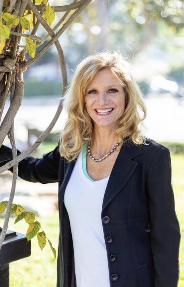 Paige Kaye smiling outdoors in a navy blazer, standing beside a vine-covered arbor